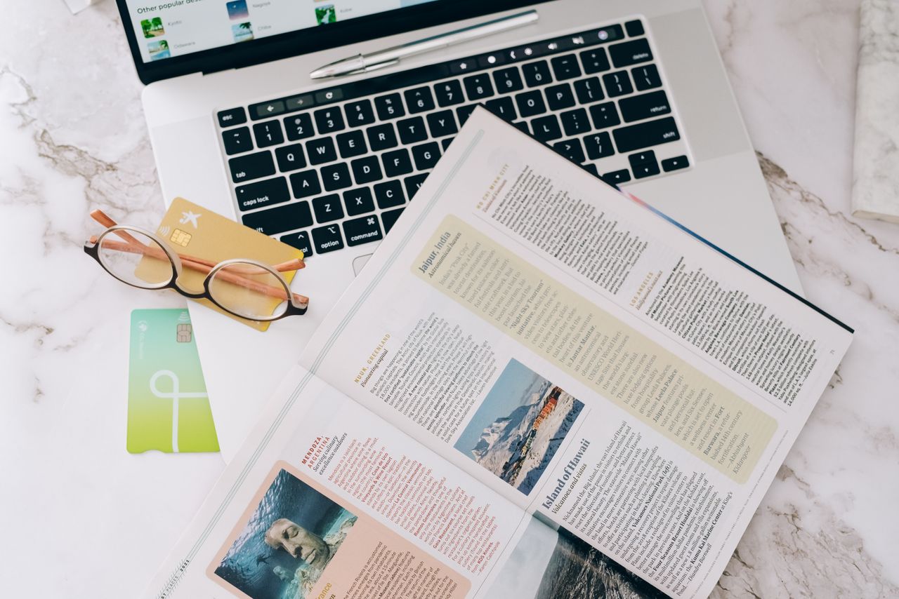 Flat lay image featuring a laptop, open magazine, credit cards, and eyeglasses on a marble table.