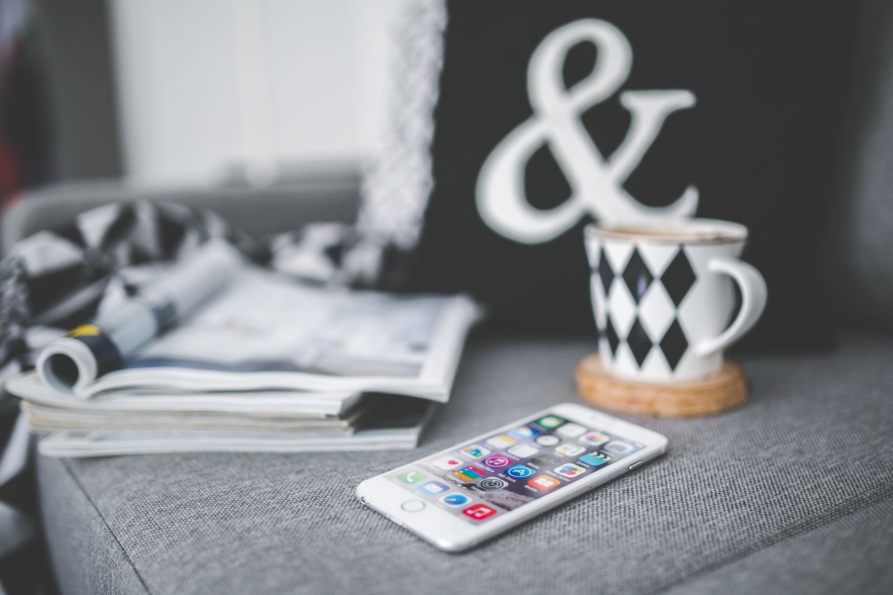 A smartphone next to a stylish coffee cup and magazines on a cozy couch setting.