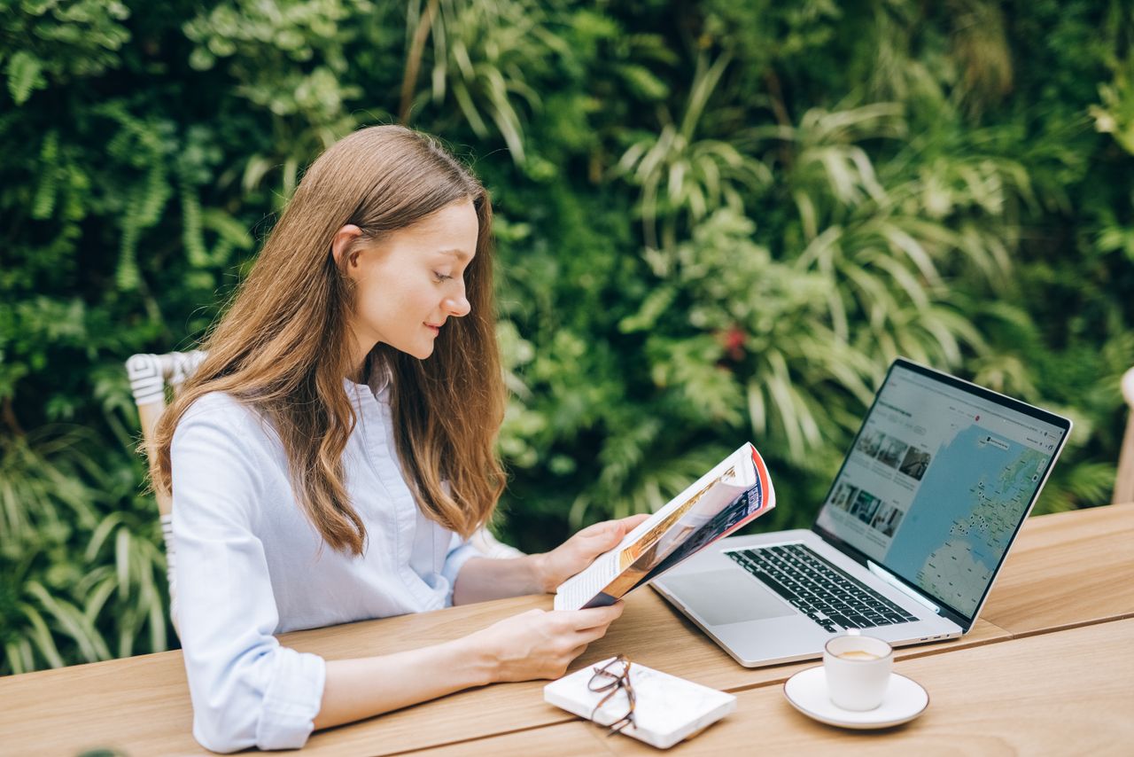 Femme caucasienne lisant un magazine à une table en bois avec un ordinateur portable, en extérieur.